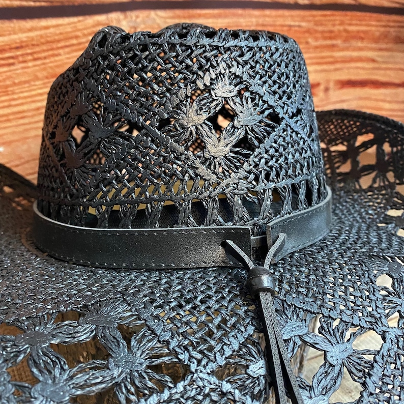 May include: A black woven straw cowboy hat with a black leather band and a black leather tassel. The hat has a wide brim and a decorative pattern.