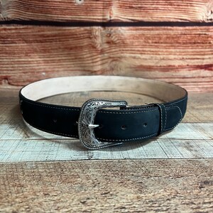 May include: A black leather belt with a silver buckle featuring an ornate design. The belt is on a wooden surface.