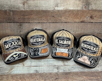 Könnte beinhalten: Vier Strohhüte mit schwarzen Netzrücken und Lederbesatz. Jeder Hut trägt einen anderen Ländernamen in einem silbernen und schwarzen Patch: Mexiko, Guatemala, El Salvador und Honduras.