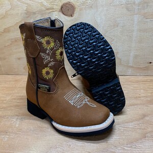 May include: Brown leather children's cowboy boots with a sunflower embroidery design. The boots have a zipper on the side and a black rubber sole. The text "CARRETA BOOTS" is on the side of the boot.