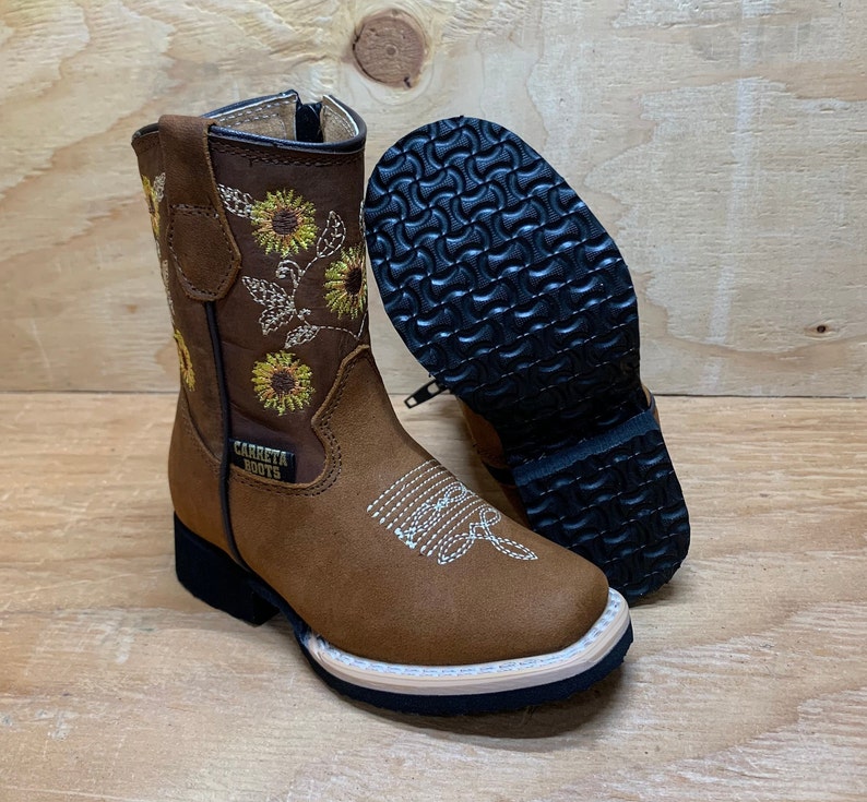 May include: Brown leather children's cowboy boots with a sunflower embroidery design. The boots have a zipper on the side and a black rubber sole. The text "CARRETA BOOTS" is on the side of the boot.