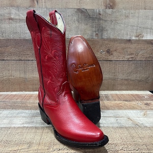 May include: A pair of red leather cowboy boots with intricate stitching and a pointed toe. The boots have a black sole and heel, and the sole of one boot is visible, showing a brand logo. The boots are set against a wooden background.