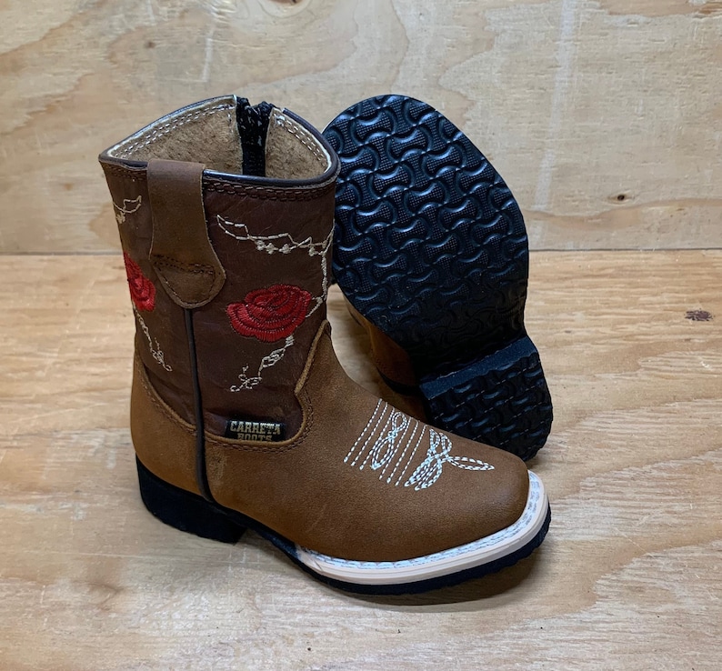 May include: Brown leather children's cowboy boots with red embroidered roses and a white stitching detail. The boots have a black rubber sole and the text "CARRETH BOOTS" is printed on the side.