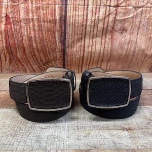 May include: Two black leather belts with silver rectangular buckles. One belt has a snakeskin pattern, and the other has a textured design. The belts are displayed on a wooden surface with a wood-paneled background.