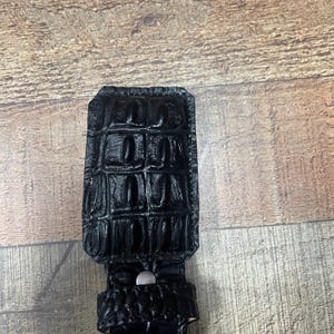 K&ouml;nnte beinhalten: Eine schwarze G&uuml;rtelschnalle in rechteckiger Form mit einer gl&auml;nzenden, strukturierten Oberfl&auml;che, die das Aussehen von Alligatorhaut nachahmt. Die Schnalle weist ein markantes, erhabenes Gittermuster auf der Oberfl&auml;che auf und enth&auml;lt einen kleinen, silberfarbenen Metallniet.