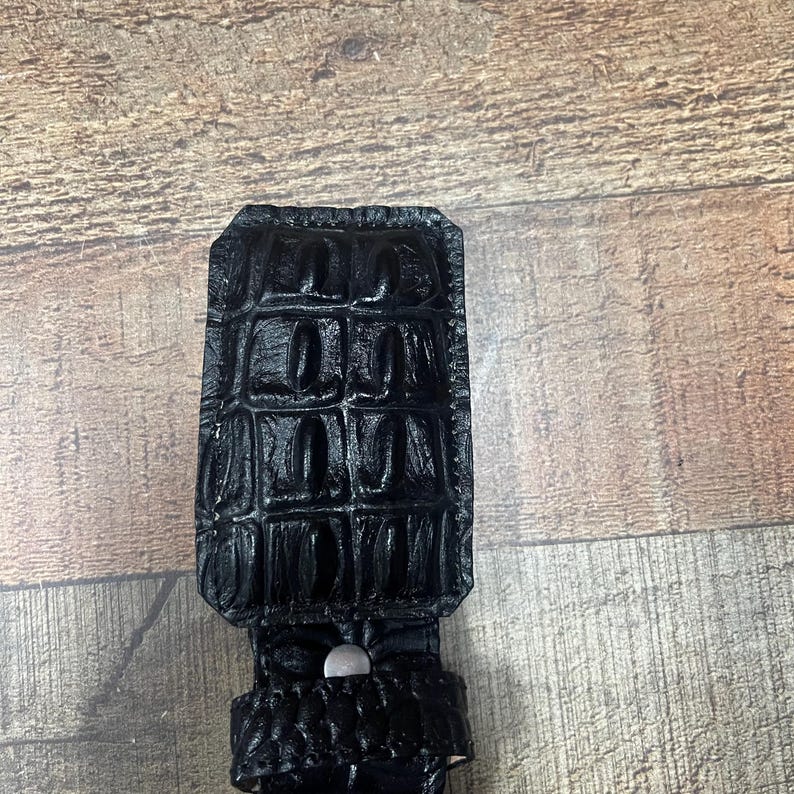K&ouml;nnte beinhalten: Eine schwarze G&uuml;rtelschnalle in rechteckiger Form mit einer gl&auml;nzenden, strukturierten Oberfl&auml;che, die das Aussehen von Alligatorhaut nachahmt. Die Schnalle weist ein markantes, erhabenes Gittermuster auf der Oberfl&auml;che auf und enth&auml;lt einen kleinen, silberfarbenen Metallniet.
