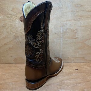 Op de afbeelding: Bruine leren cowboylaarzen met bloemenborduurwerk in gouden en bruine draad. De laarzen hebben een spitse neus en een lage hak.