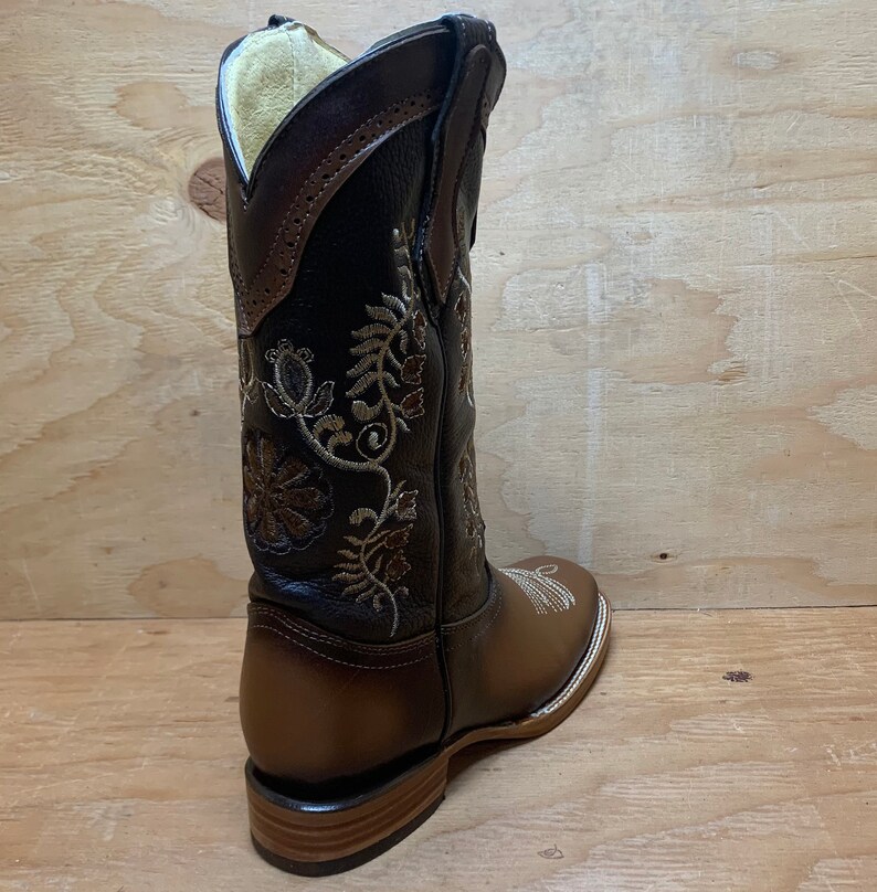 Op de afbeelding: Bruine leren cowboylaarzen met bloemenborduurwerk in gouden en bruine draad. De laarzen hebben een spitse neus en een lage hak.