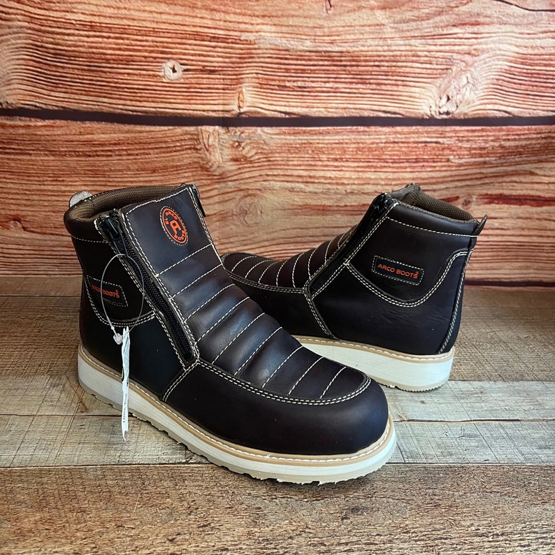 May include: A pair of brown leather boots with a zipper on the side. The boots have a white sole and a black stitching detail. The boots have the text "ARCO BOOTS" on the side.