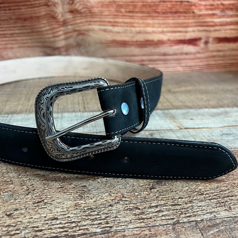May include: A black leather belt with a silver buckle featuring an ornate design. The belt has a stitched edge and a smooth finish.