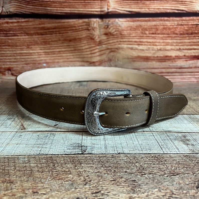 May include: A brown leather belt with a silver buckle featuring an ornate design. The belt is on a wooden surface.