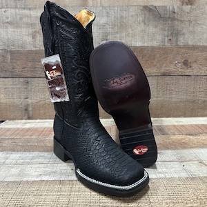 May include: Black leather cowboy boots with a textured pattern and square toe. One boot is upright, the other is lying on its side, revealing the sole. The boots have a white stitched border and a tag attached.