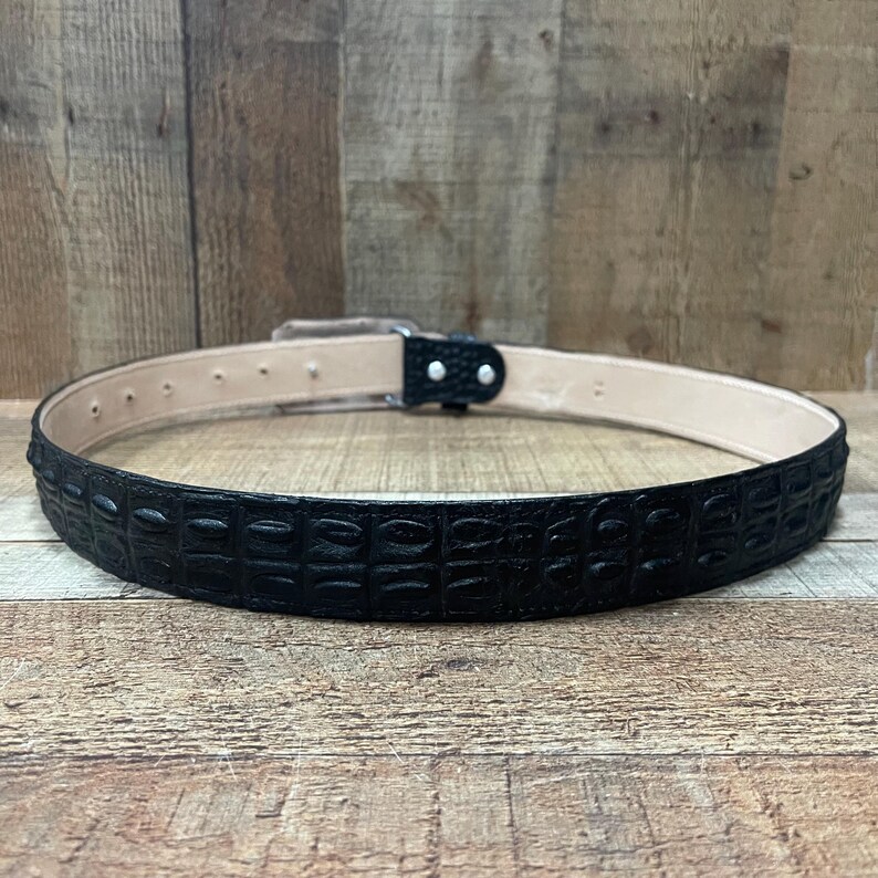 K&ouml;nnte beinhalten: Ein schwarzer G&uuml;rtel mit einer texturierten Krokodilpr&auml;gung auf der Au&szlig;enseite und einem glatten beigen Innenfutter. Der G&uuml;rtel verf&uuml;gt &uuml;ber eine schwarze Schnalle mit zwei silberfarbenen Metallnieten und eine Reihe von Verstelll&ouml;chern.