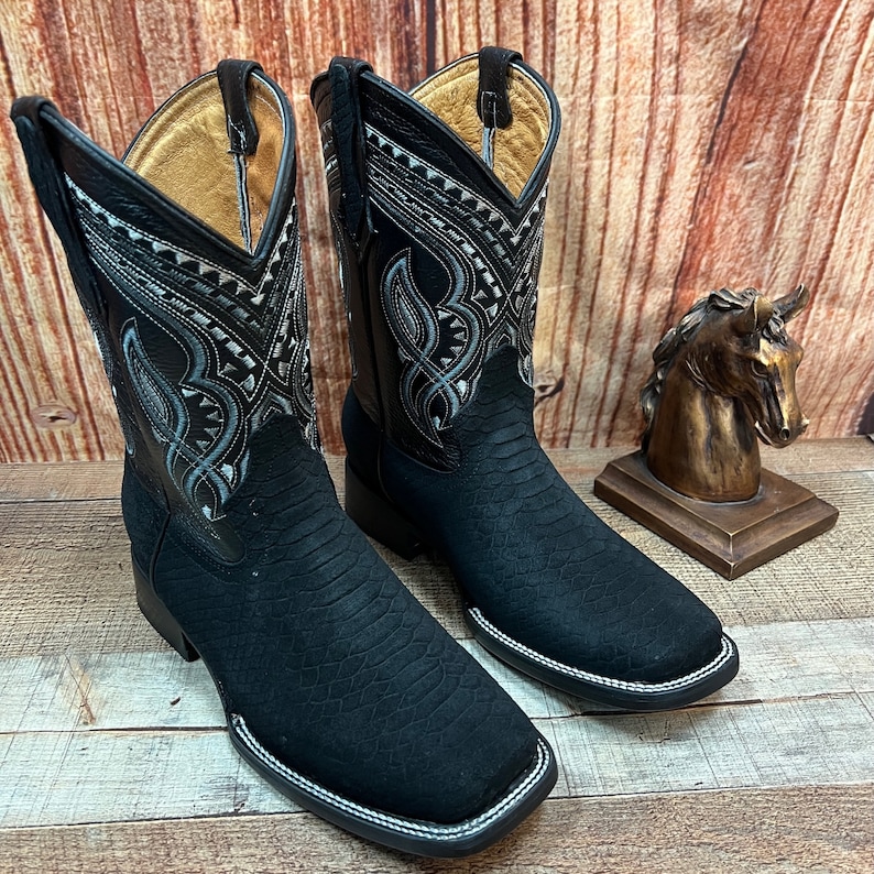 May include: A pair of black cowboy boots with a snakeskin pattern and intricate silver embroidery on the shaft. The boots have a pointed toe and a white stitching detail around the sole.