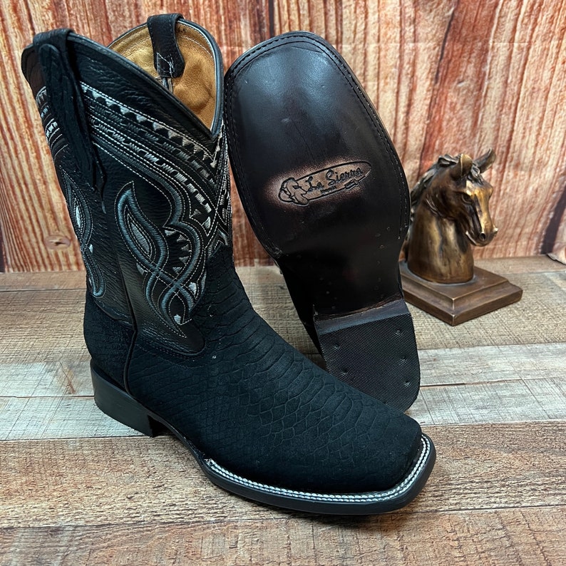 May include: Black leather cowboy boots with a decorative silver design on the shaft. The boots have a pointed toe and a stacked heel. The boots are made by La Sierra.