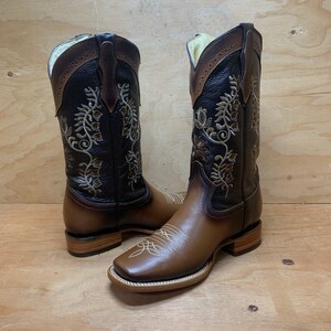 Op de afbeelding: Een paar bruine leren cowboylaarzen met ingewikkelde bloemenborduursels in een lichtere bruine kleur. De laarzen hebben een vierkante neus en een lage hak.