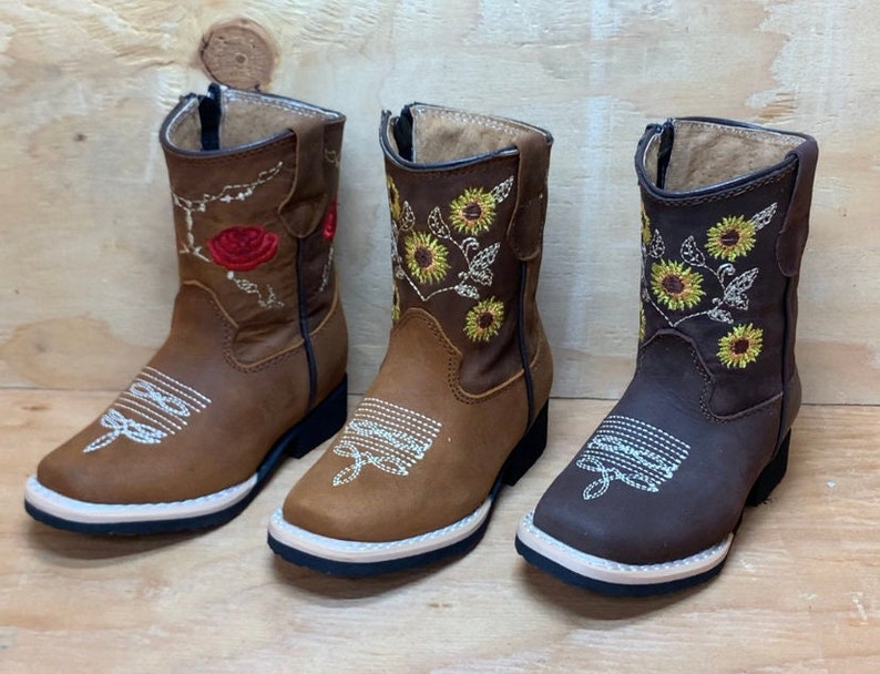 May include: Three pairs of brown leather children's cowboy boots with embroidered floral designs. One pair features red roses, while the other two pairs feature yellow sunflowers.