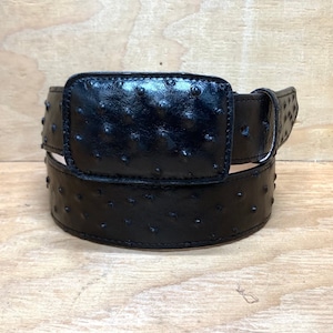 Op de afbeelding: Een zwarte riem van struisvogelhuid met een rechthoekige gesp. De riem is gemaakt van hoogwaardig leer en heeft een klassiek ontwerp.