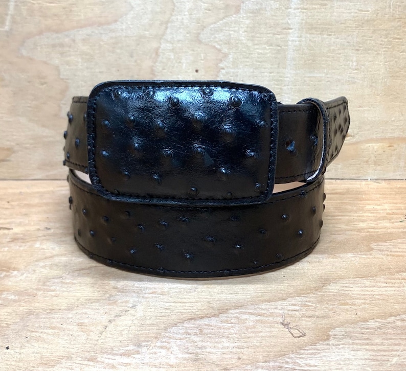 Op de afbeelding: Een zwarte riem van struisvogelhuid met een rechthoekige gesp. De riem is gemaakt van hoogwaardig leer en heeft een klassiek ontwerp.