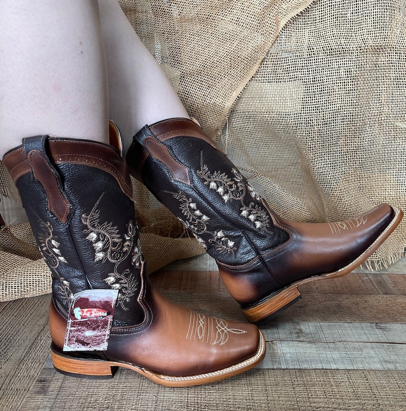Op de afbeelding: Een paar bruine en beige leren cowboylaarzen met bloemenborduurwerk. De laarzen hebben een spitse neus en een lage hak.