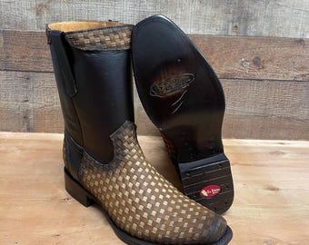 Könnte beinhalten: Ein Paar braune und schwarze Leder-Cowboystiefel. Die Stiefel haben ein gewebtes Design am Fuß und einen einfarbigen schwarzen Schaft. Die Sohle eines Stiefels ist sichtbar und zeigt den Markennamen und das Logo. Die Stiefel werden auf einer Holzoberfläche präsentiert.