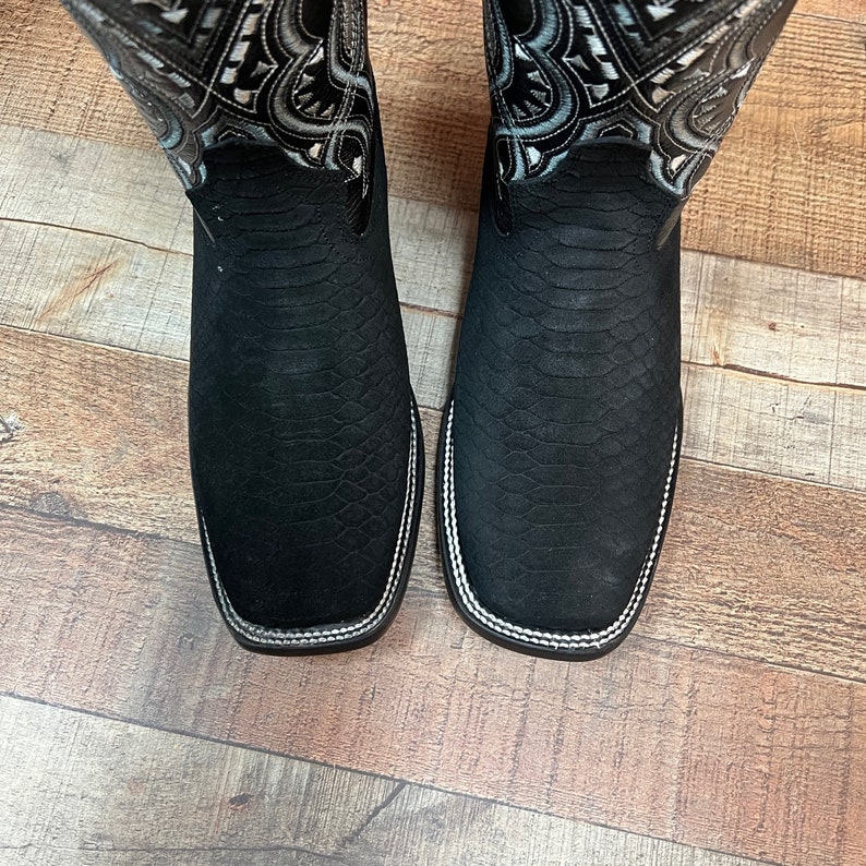 May include: A pair of black cowboy boots with a snakeskin pattern and white stitching. The boots have a decorative design on the top of the shaft.