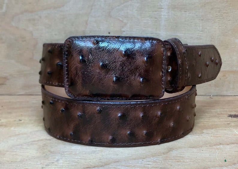 Op de afbeelding: Een bruine leren riem met een struisvogelhuidpatroon en een bijpassende gesp. De riem is dubbelgevouwen en de gesp is zichtbaar.