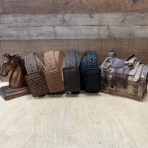 May include: Five woven leather belts in different shades of brown and black. The belts are displayed on a wooden surface with a miniature horse head and a saddle on a chest to the right.