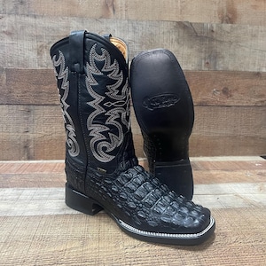 May include: Black cowboy boots with a square toe and a crocodile-like texture on the foot. The shaft features intricate white stitching. The sole is black, and the boots are displayed on a wooden surface.
