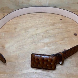 Op de afbeelding: Een bruine leren riem met een struisvogelhuidpatroon en een bijpassende gesp. De riem ligt plat op een houten oppervlak.