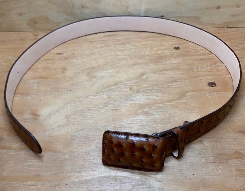 Op de afbeelding: Een bruine leren riem met een struisvogelhuidpatroon en een bijpassende gesp. De riem ligt plat op een houten oppervlak.