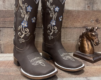 May include: A pair of brown leather cowboy boots with floral embroidery in blue and gold. The boots have a square toe and a white sole. A bronze horse statue is in the background.