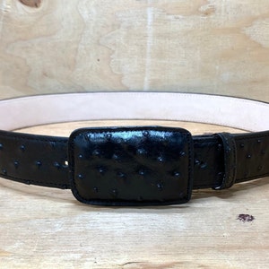 Op de afbeelding: Een zwarte leren riem met een struisvogelhuidpatroon en een vierkante gesp. De riem is gemaakt van hoogwaardig leer en is ontworpen voor duurzaamheid en stijl.
