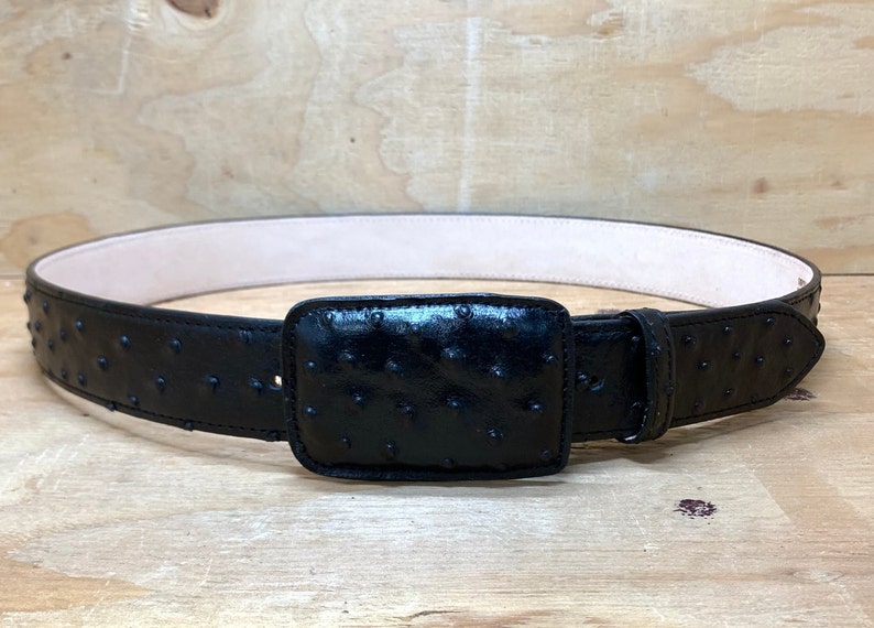 Op de afbeelding: Een zwarte leren riem met een struisvogelhuidpatroon en een vierkante gesp. De riem is gemaakt van hoogwaardig leer en is ontworpen voor duurzaamheid en stijl.