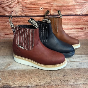 May include: Three pairs of brown leather ankle boots with white soles. The boots have a pull-on design and a stitched detail around the ankle. The boots are labeled "Carreta Boots" on the side.