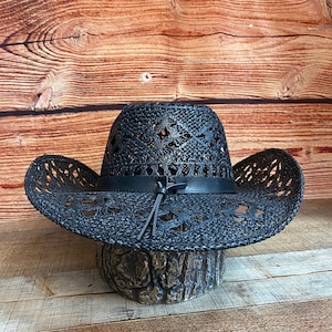 May include: A black woven straw cowboy hat with a leather band and a braided leather tie. The hat has a decorative cut-out pattern.