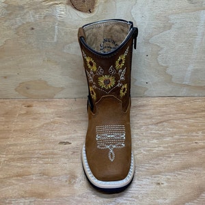 May include: Brown leather children's boots with a zipper on the side. The boots have a sunflower embroidery design on the front and back. The boots are made of genuine leather and have a rubber sole.
