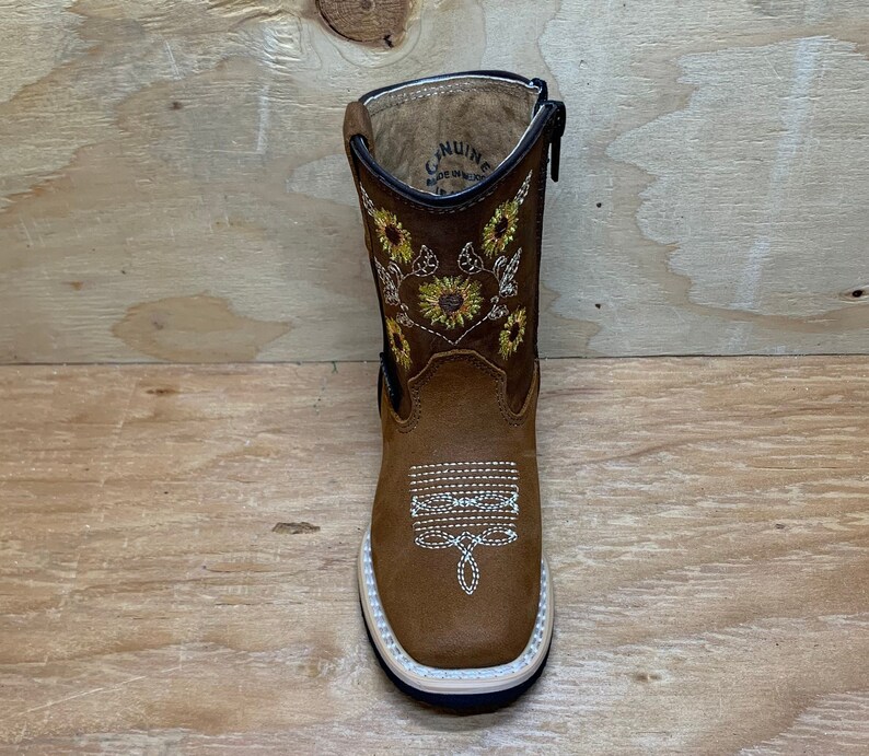 May include: Brown leather children's boots with a zipper on the side. The boots have a sunflower embroidery design on the front and back. The boots are made of genuine leather and have a rubber sole.