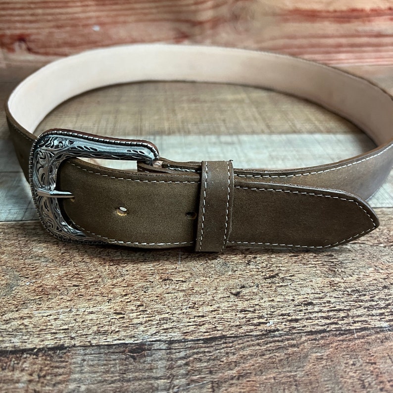May include: A brown leather belt with a silver buckle featuring an engraved design. The belt has a stitched edge.