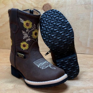 May include: Brown leather children's cowboy boots with a zipper on the side. The boots have a sunflower embroidery design and the text "Carreta Boots" on the side.