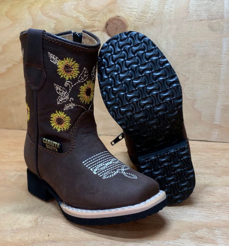May include: Brown leather children's cowboy boots with a zipper on the side. The boots have a sunflower embroidery design and the text "Carreta Boots" on the side.