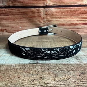 May include: Black leather belt with a silver buckle and white embroidery. The embroidery features a floral design.