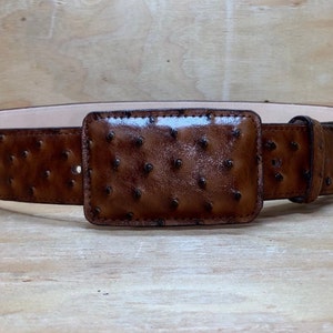 Op de afbeelding: Een bruine leren riem met een struisvogelhuidpatroon en een rechthoekige gesp. De riem is gemaakt van hoogwaardig leer en heeft een klassiek ontwerp.