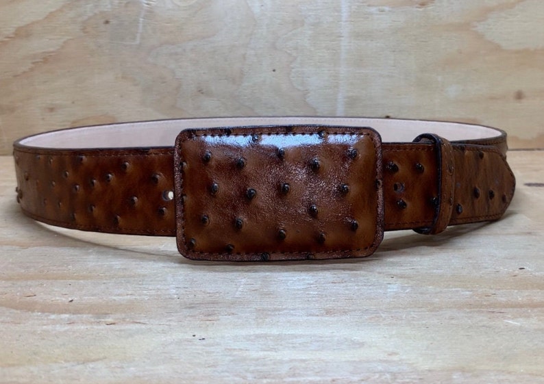 Op de afbeelding: Een bruine leren riem met een struisvogelhuidpatroon en een rechthoekige gesp. De riem is gemaakt van hoogwaardig leer en heeft een klassiek ontwerp.