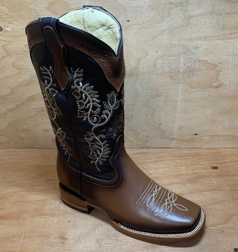 Op de afbeelding: Bruine leren cowboylaarzen met bloemenborduurwerk. De laarzen hebben een vierkante neus en een lage hak.