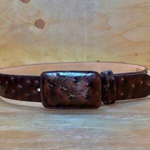 Op de afbeelding: Een bruine leren riem met een struisvogelprint en een rechthoekige gesp. De riem heeft een rij kleine studs langs de randen.