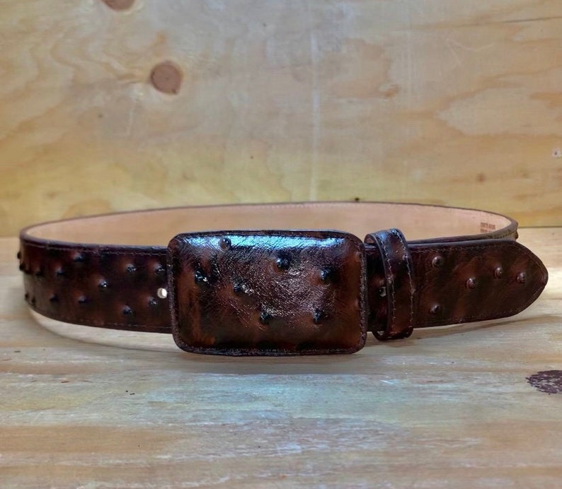 Op de afbeelding: Een bruine leren riem met een struisvogelprint en een rechthoekige gesp. De riem heeft een rij kleine studs langs de randen.