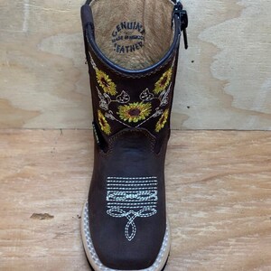 May include: Brown leather children's cowboy boots with a side zipper and sunflower embroidery. The boots have a white stitching detail and the words "Genuine Leather Made in Mexico" are printed on the inside of the boot.