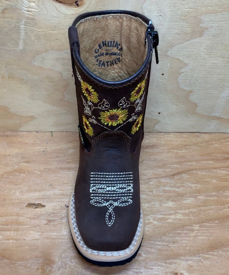 May include: Brown leather children's cowboy boots with a side zipper and sunflower embroidery. The boots have a white stitching detail and the words "Genuine Leather Made in Mexico" are printed on the inside of the boot.