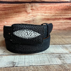 May include: A black belt with a textured surface and a rectangular buckle. The buckle features a white and gray design in the center. The belt is made of a durable material and is suitable for everyday wear.